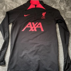 Liverpool Nike långärmad träningströja S - Säljer en svart och röd Liverpool FC träningströja från Nike i Dri-Fit material. Tröjan har lång ärm, halv dragkedja och tryck med AXA, Nike-logga och LFC-märke på bröstet. Slim fit-modell, perfekt för fotbollsträning eller support. Skicket är 9,5/10 använd fåtal gånger. 
