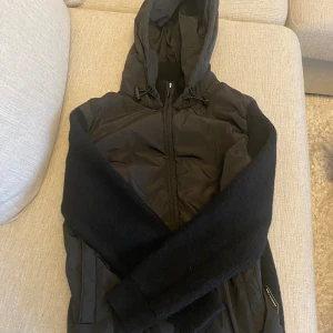 Cardigan Jacka - Svart hybridjacka från Porsche BOSS i storlek M. Jackan har huva, dragkedja och kombinerar stickade partier med vadderade paneler. Den är perfekt i varma vinterdagar och perfekt i hösten då skicket är 10/10 helt ny och oanvänd.