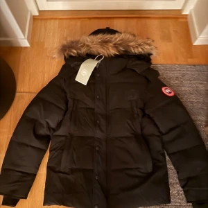 Svart dunjacka från Canada Goose - Stk S men passar även XS. Pris kan diskuteras!! Säljer en svart dunjacka från Canada Goose med huva och snygg fuskpäls runt kanten. Jackan har dragkedja och knappar framtill, samt klassisk logga på ärmen. Perfekt för kalla vinterdagar och riktigt varm. Flera praktiska fickor och stilren design.