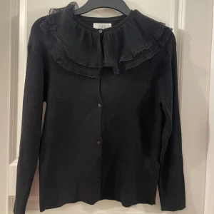 Svart kofta med volangkrage från Zara - Supersnygg svart kofta från Zara med ribbad struktur och stora volanger runt kragen. Långa ärmar och knäppning framtill. Perfekt för dig som vill ha något extra till din outfit!