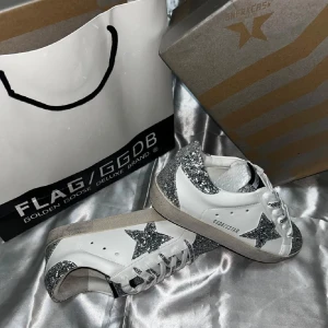 Golden Goose sneakers med glitterdetaljer - Golden Goose sneakers i vitt skinn med silverglittriga detaljer på tå, häl och stjärna på sidan. Klassisk låg modell med snörning och platt sula. Märkets logga syns på plösen och sidan. Perfekta för dig som vill sticka ut med lite extra bling.
