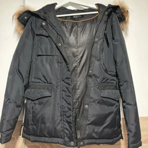 Svart parkajacka med äkta päls från Hollies - Svart parkajacka från Hollies med huva och äkta päls. Jackan har flera praktiska fickor framtill, dragkedja och knappar. Perfekt för kyliga dagar med sin varma och skyddande design. Klassisk och stilren look.