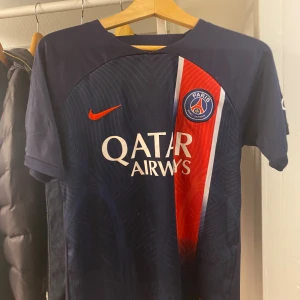 PSG matchtröja Nike blå/röd - Paris Saint-Germain fotbollströja från Nike i mörkblått med röd och vit rand på framsidan. Tröjan har klubbmärke, Qatar Airways-tryck och korta ärmar. Tillverkad i lätt och ventilerande material, perfekt för match eller träning.