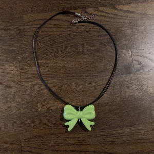 Halsband med grön rosett - Ett svart halsband med ett stort limegrönt rosett-hänge i plast. Halsbandet har justerbar kedja och karbinhake i silverfärg. Perfekt statement-piece för dig som gillar färg och popp av form i din stil.