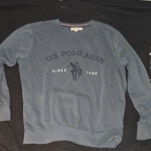 Blå sweatshirt från U.S. Polo Assn. - Blå sweatshirt från U.S. Polo Assn. med tryckt logga och polospelare på bröstet. Tröjan har rund halsringning, långa ärmar och ribbade muddar vid ärmslut och nederkant. Perfekt för en avslappnad och sportig stil.