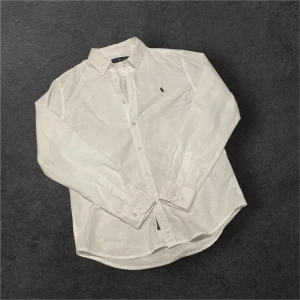 Ralph Lauren skjorta - Klassisk vit skjorta från Ralph Lauren i linne med broderad marinblå logga på bröstet. Skjortan har knappar framtill, lång ärm med manschett och tidlös krage. Perfekt för en clean och stilren look.