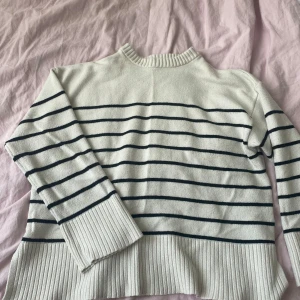 Vit stickad randig tröja med slits - Säljer en vit stickad tröja med svarta ränder. Tröjan har långärmade ribbade muddar och bred ribbad nederkant med slits på sidorna. Perfekt för dig som gillar klassisk stil med en twist. Rund halsringning och mjukt material.