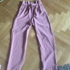 Rosa kostymbyxor med bälte från Bik Bok - Snygga ljusrosa kostymbyxor från Bik Bok i storlek XS. Byxorna har hög midja, avsmalnande ben och ett matchande tygbälte i midjan. Tillverkade i ett mjukt, vävt material med hällor och knäppning framtill. Perfekta för en trendig och chic look.