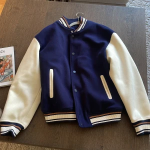 Blå och vit bomberjacka - Zara - Bomberjacka från Zara i mörkblått med vita ärmar och ribbade muddar med blå, vita och bruna ränder. Jackan har quiltat foder, tryckknappar och två fickor fram. 