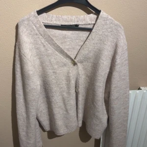 Beige stickad kofta med knappar - Mysig beige stickad kofta med v ringning och tre knappar framtill. Koftan har en croppad passform och långa ärmar, perfekt för lager på lager under kyliga dagar.