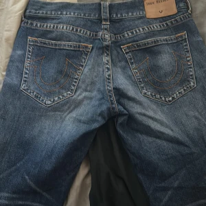 Blå jeans från True Religion, stl 34 - Säljer ett par klassiska blå jeans från True Religion i storlek 34. Jeansen har tydliga kontrastsömmar, ikoniska bakfickor med broderad logga och snygg tvättad finish. Tillverkade i slitstarkt denim med fem fickor och knappgylf.