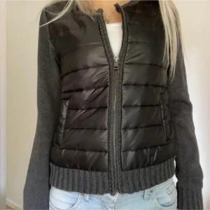 Grå pufferjacka med stickade ärmar cardigan  - Cardigan Säljer en grå pufferjacka f med quiltad front i glansigt material och stickade ärmar samt muddar. Jackan har dragkedja framtill och två fickor. Perfekt för dig som vill ha en chill och snygg look till hösten eller vintern.