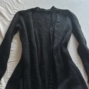 Svart stickad kofta med lång ärm - Mysig svart stickad kofta med lång ärm och öppen framtill. Koftan har ribbade muddar vid ärmslut och nederkant, perfekt att slänga på sig när det är kyligt. Enkel och stilren design som passar till det mesta.