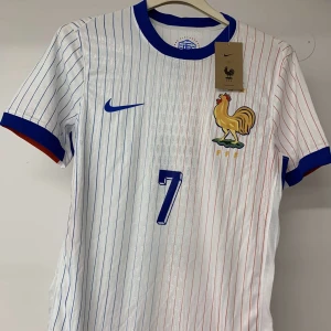 Frankrike Griezmann #7 fotbollströja Nike - Snygg vit fotbollströja från Nike med tunna blå och röda ränder, blå krage och ärmslut med röda detaljer. Franska landslagets emblem med tupp på bröstet och Griezmanns namn samt nummer 7 på ryggen. Tillverkad i lätt och ventilerande polyester.