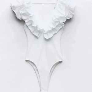 Vit body med volangdetaljer - Supersnygg vit body med djup v-ringning och stora volanger längs axlar och urringning. Perfekt för dig som vill sticka ut på stranden med en feminin och trendig look. Hög benskärning ger en modern siluett.