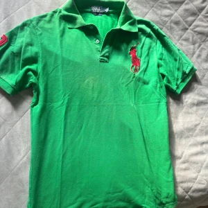 Grön pikétröja Polo Ralph Lauren XL - Snygg grön pikétröja från Polo Ralph Lauren i klassisk modell med korta ärmar. Stor röd broderad logga på bröstet och röd siffra 3 på ärmen. Tröjan har krage och knäppning framtill. Tillverkad i bomull för en skön känsla.