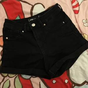Svarta jeansshorts från FB Sister - Svarta jeansshorts från FB Sister i storlek S. Klassisk femficksmodell med hög midja, knapp och dragkedja framtill. Shortsens ben är uppvikta för en chill look. Perfekta för varma dagar och enkla att matcha med allt.