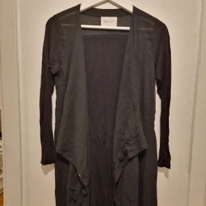 Svart lång kofta från Capri Collection - Svart lång kofta/kaftan från Capri Collection med öppen front och snyggt draperade kanter. Tunn och lätt i materialet, perfekt att slänga över en topp. Passar till många olika outfits. Skrynklig på bilden då den har endast legat I garderoben tyvärr.