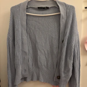 Ljusblå stickad kofta från 157 - Säljer en ljusblå stickad kofta från 157 med stora bruna knappar framtill. Koftan har lång ärm, ribbade muddar och en avslappnad passform. Perfekt att slänga över en t-shirt när det är lite kyligt ute.