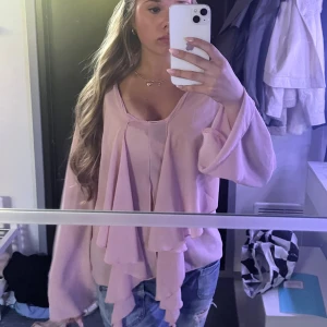 Ljusrosa blus med volang och knyt - Supersöt ljusrosa blus med långa, vida ärmar och v-ringning. Framtill har den en stor volangdetalj som kan knytas till en rosett eller hänga löst. Tunt, flowy material som ger en romantisk vibe och passar perfekt till jeans.