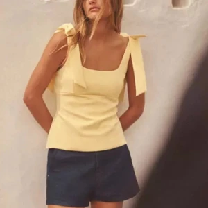 Gul linnetopp med rosetter på axlarna - Supersöt ljusgul linnetopp med breda axelband och fina rosetter på axlarna. Toppen har en fyrkantig ringning och figurnära passform som ger en fräsch och somrig vibe. Perfekt att matcha med shorts eller kjol för en chill look. 