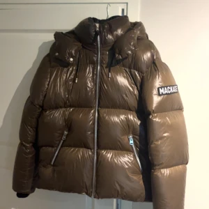 Brun pufferjacka från Mackage - Säljer en brun pufferjacka från Mackage med glansig finish och stor huva. Jackan har dragkedja framtill, två snedställda fickor med dragkedja och tydlig logga på ärmen. Perfekt för kalla dagar när du vill hålla stilen och värmen.