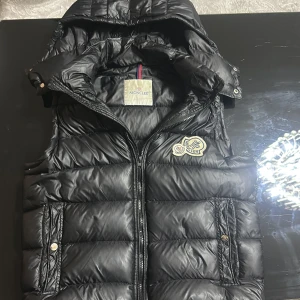 Svart dunväst från Moncler - Svart dunväst från Moncler med quiltad design och huva. Västen har dragkedja framtill, två fickor med knappstängning och Moncler-logga på bröstet. Materialet är polyester och dun, vilket gör den både lätt och varm. Perfekt för kyliga dagar.