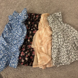 Fyra korta sommarkjolar i olika mönster - Fyra superfina kortklänningar i olika färger och mönster. En blå med vita blommor, en svart med röda och beige blommor, en beige med vågigt mönster och volang, samt en grå med vita blommor. Perfekta för varma dagar och ger en fräsch vibe. Allla för 100kr eller 40kr/st
