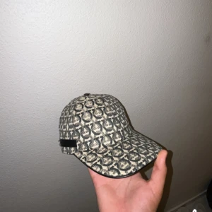 Salvatore Ferragamo mönstrad keps - Snygg keps från Salvatore Ferragamo med grått och beige mönster över hela kepsen. Justerbar rem baktill med metallspänne och svart läderdetalj med logga på sidan. Tillverkad i bomull och polyester, perfekt för att lyfta din outfit.
