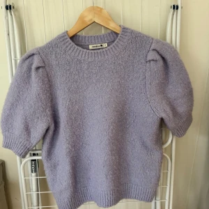 Ljuslila stickad tröja från Indiska -  ljuslila stickad tröja från Indiska med puffiga korta ärmar och rund halsringning. Tröjan har en mjuk och mysig känsla, perfekt för lager på lager under höst och vår. Snygg ribbad kant vid ärmslut och nederkant.