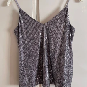 Glittrig silver linnetopp från Amisu - Säljer en glittrig silverfärgad linnetopp från Amisu med tunna axelband och v-ringning. Toppen är täckt av paljetter som ger ett riktigt snyggt partyvibe. Perfekt för dig som vill sticka ut och glänsa lite extra. Sitter snyggt och är lätt att matcha med både jeans och kjol.
