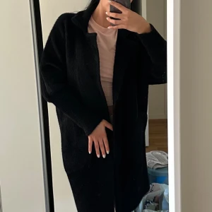 Svart ”alpacka” kappa med fickor - Lång svart alpacka kappa med krage och stora fickor framtill. Jackan har en oversized passform och stängs med en knapp. Mjuk och fluffig känsla, perfekt för kyligare dagar.