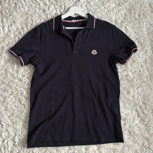 Mörkblå Moncler pikétröja med logga - Mörkblå pikétröja från Moncler med klassisk krage och korta ärmar. Tröjan har Moncler-logga på bröstet och snygga rödvita detaljer på krage och ärmslut. Tillverkad i bomull för en skön och stilren känsla.