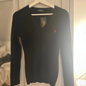 Svart stickad tröja Polo Ralph Lauren - Svart stickad tröja från Polo Ralph Lauren med v-ringning och klassisk röd logga broderad på bröstet. Tröjan har lång ärm och en slim passform som sitter snyggt. Perfekt för dig som gillar stilrena och tidlösa plagg. M men sitter som s/xs