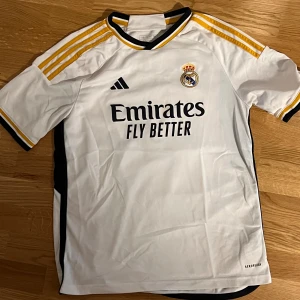 Real Madrid Arda Güler matchtröja Adidas - Real Madrid fotbollströja med Arda Güler och nummer 24 på ryggen. Vit med svarta och gula detaljer, klassiska Adidas-ränder på axlarna och klubbmärke på bröstet. Tillverkad i lätt och ventilerande polyester med AEROREADY-teknologi.