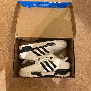 Adidas Rivalry Low vita sneakers - Adidas Rivalry Low sneakers i vitt med svarta detaljer och klassiska tre ränder på sidan. Skorna har snörning, platt sula och rund tå. Tillverkade i syntetmaterial och skinn för en clean och sportig look. Perfekta för dig som gillar retrovibbar och streetstyle. Pris kan diskuteras!