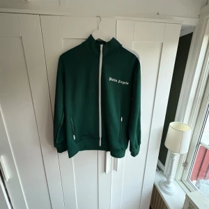 Grön Palm Angels zip hoodie - Snygg mörkgrön hoodie från Palm Angels med vit dragkedja och vita ränder längs ärmarna. Tröjan har hög krage, två fickor med dragkedja och logotypen broderad på bröstet. Perfekt för dig som gillar streetwear och sportig stil.