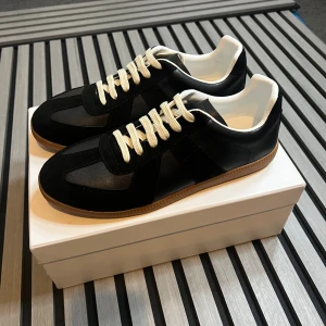 Maison Margiela gats - Maison Margiela sneakers i svart skinn och mocka med vita snören och vit insida. Klassisk gummisula i beige och diskret branding på insidan. Snygg och tidlös modell som passar till allt. Perfekt för dig som gillar minimalistisk stil.