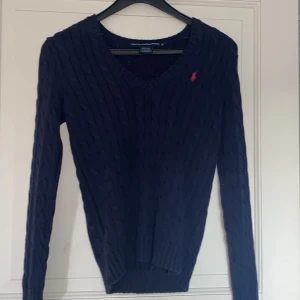 Mörkblå stickad tröja Ralph Lauren - Mörkblå kabelstickad tröja från Ralph Lauren Sport med röd broderad logga på bröstet. V-ringad modell med långa ärmar och ribbade muddar. Perfekt för lager-på-lager och snygg till jeans eller kjol.