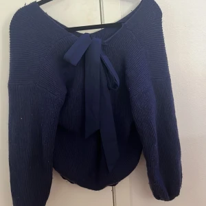 Mörkblå stickad tröja med rosett - Mörkblå stickad tröja med långärmad design och bred v-ringning. Baksidan har en snygg rosettdetalj i matchande tyg som ger en cool touch. Tröjan har ribbad struktur och är lite loose i passformen, perfekt för dig som gillar en chill och stilren look. Dock ett litet sträck i tyget som man ser på sista bilden