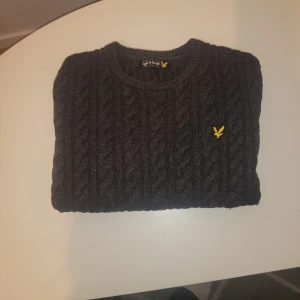 Mörkgrå stickad tröja från Lyle & Scott - Säljer en mörkgrå stickad tröja från Lyle & Scott med klassiskt kabelstickat mönster och rund hals. Tröjan har en gul broderad logga på bröstet och är perfekt för kyliga dagar.
