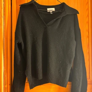 Svart stickad tröja från AWARE XS - Svart stickad tröja från AWARE i storlek XS. Tröjan har en klassisk krage, v-ringning och långa ärmar. Ribbstickade muddar vid ärmslut och nederkant ger en snygg detalj. Perfekt för lager-på-lager under kyliga dagar.