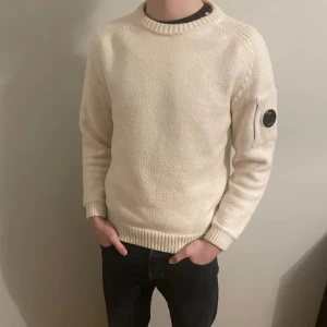 Vit stickad tröja från C.P. Company - Säljer en stilren vit stickad tröja från C.P. Company. Tröjan har rund halsringning, ribbade muddar och en ikonisk svart linsdetalj på ärmen. Perfekt för dig som gillar clean och modern streetwear.