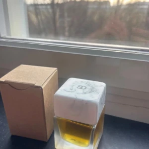 Sora Dora Jany - Säljer en stilren parfym från JANY med en fyrkantig glasflaska och marmorerad vit kork. Flaskan har en transparent design som visar den gyllene vätskan inuti. Kommer med en enkel brun kartongförpackning. Perfekt för dig som gillar minimalistisk och modern estetik. Online kostar denna parfym 2500-4000kr. Parfymen är endast testad
