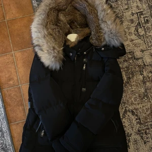 Svart dunjacka med päls från Cedrico - Säljer en svart dunjacka från Cedrico i storlek L med stor fluffig päls på huvan. Jackan har flera fickor med dragkedja och tryckknappar, samt snygga detaljer i skinn. Perfekt för kalla vinterdagar och har en riktigt schysst look.