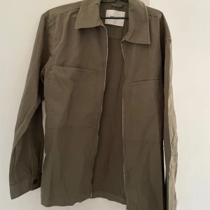 Olivgrön overshirt från NN.07 - Säljer en stilren olivgrön overshirt från NN.07 i ekologisk bomull. Jackan har raka linjer, två stora bröstfickor, klassisk krage och dragkedja framtill. Perfekt att slänga på över en t-shirt för en clean look.