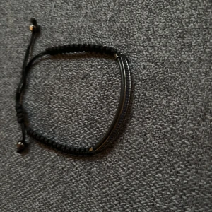 Svart justerbart armband från bvaldi - Snyggt svart armband med flätad textil och en böjd metallstav framtill från Bvaldi. Armbandet har justerbar knytning med små metallpärlor i ändarna. Perfekt accessoar för en stilren look.
