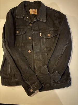 Svart jeansjacka från Levi's, strl 36 - Klassisk svart jeansjacka från Levi's i 100% bomull. Jackan har knappar framtill, två bröstfickor med lock och knapp samt två sidofickor. Snygg och tidlös modell med raka linjer och krage. Perfekt för dig som gillar en clean och stilren look.