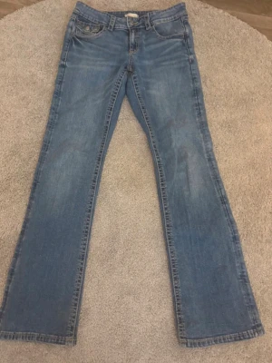 Blå bootcut jeans från Gina Tricot - Säljer ett par klassiska blå jeans från Gina Tricot med bootcut passform. Jeansen har snygga kontrastsömmar, fem fickor och knappstängning fram. Materialet är slitstarkt denim i bomull och bakfickorna har lock med knappdetalj.