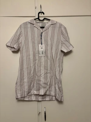 Randig kortärmad skjorta Larsson & Co - Kortärmad skjorta från Larsson & Co i vitt med tunna bruna ränder. Skjortan har klassisk krage, knäppning framtill och är tillverkad i bomull. Perfekt för en avslappnad och stilren look.